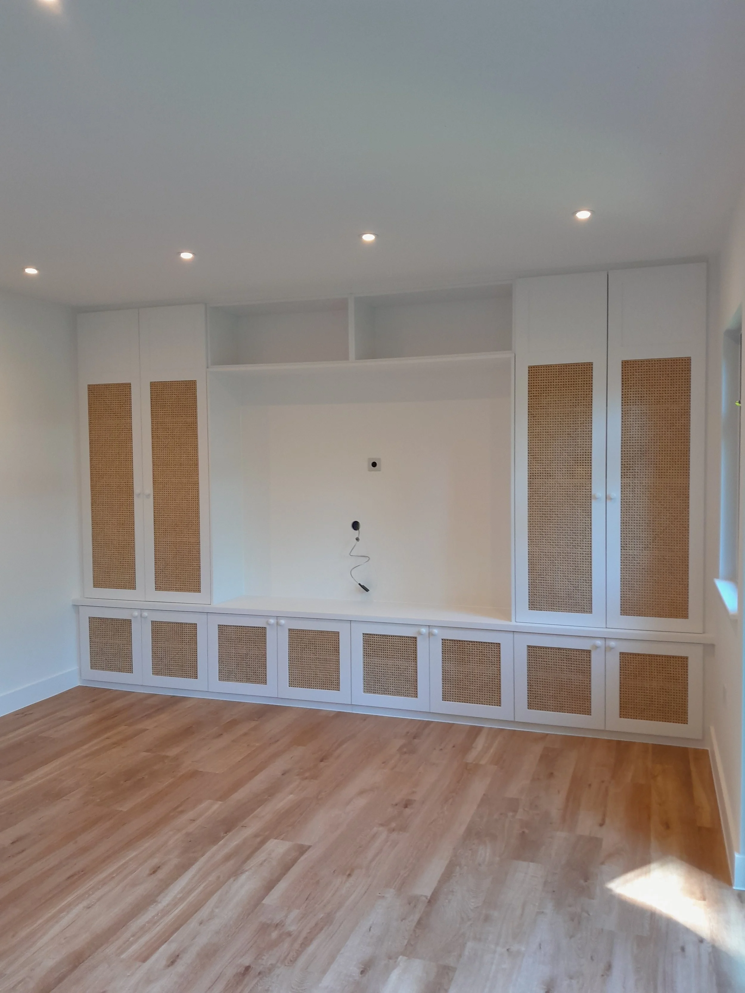 Empty built-in white cabinet with woven rattan panels, in a room with light-colored hardwood floors and a white ceiling with recessed lighting.