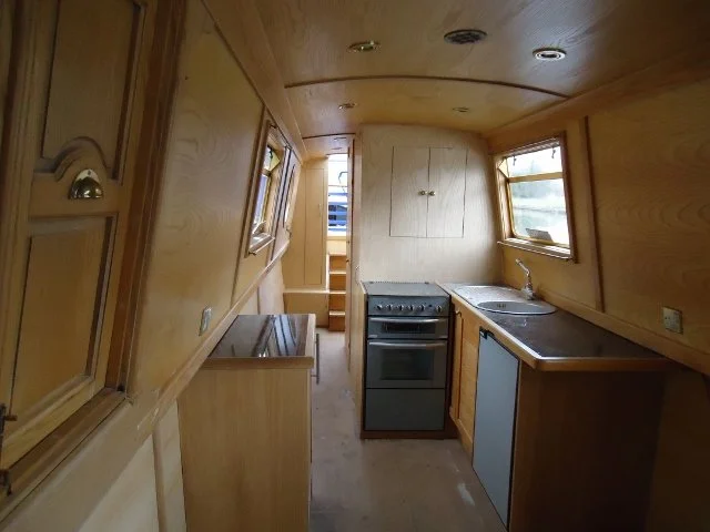 Small wooden kitchen with two windows, stove, sink, and limited counter space inside a boat or tiny home.