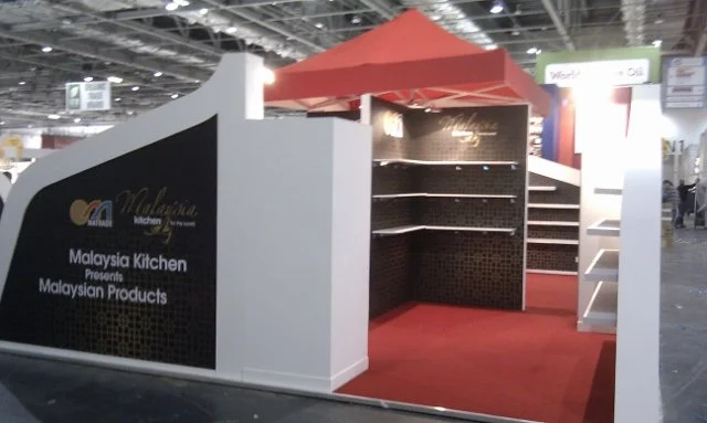 Trade show booth for Malaysia Kitchen showcasing Malaysian products, featuring black and white panels with red canopy in an exhibition hall.