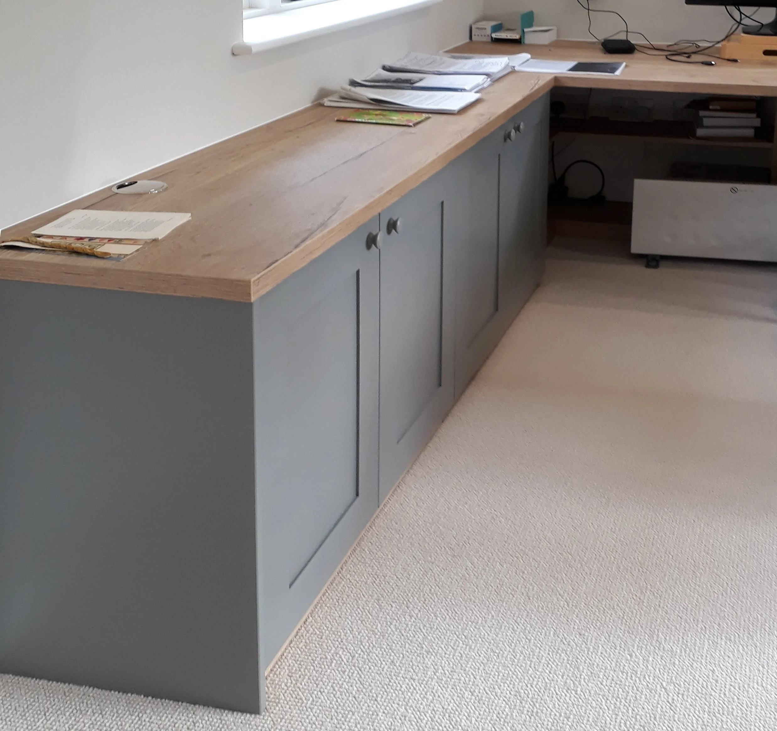 A wooden desk with gray cabinet doors, cluttered with papers and documents near a window, in a room with beige carpet.