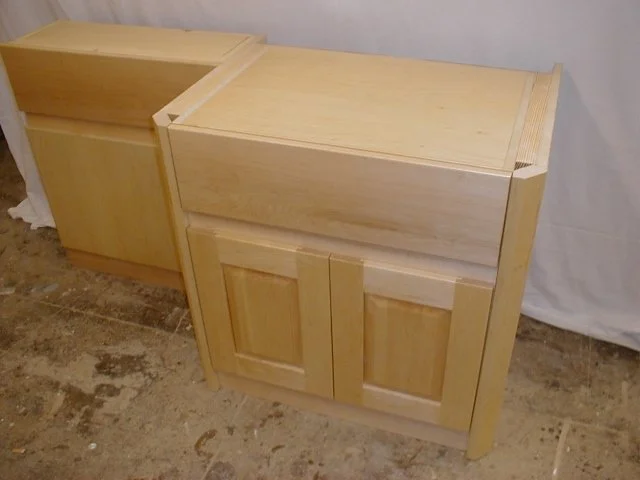 Two unfinished wooden cabinets with double doors, placed on a rough concrete floor.