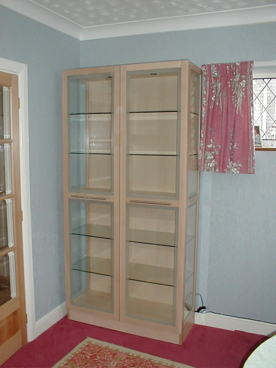 Empty display cabinet with glass doors and shelves in a room with curtains, a window, and a floral area rug.