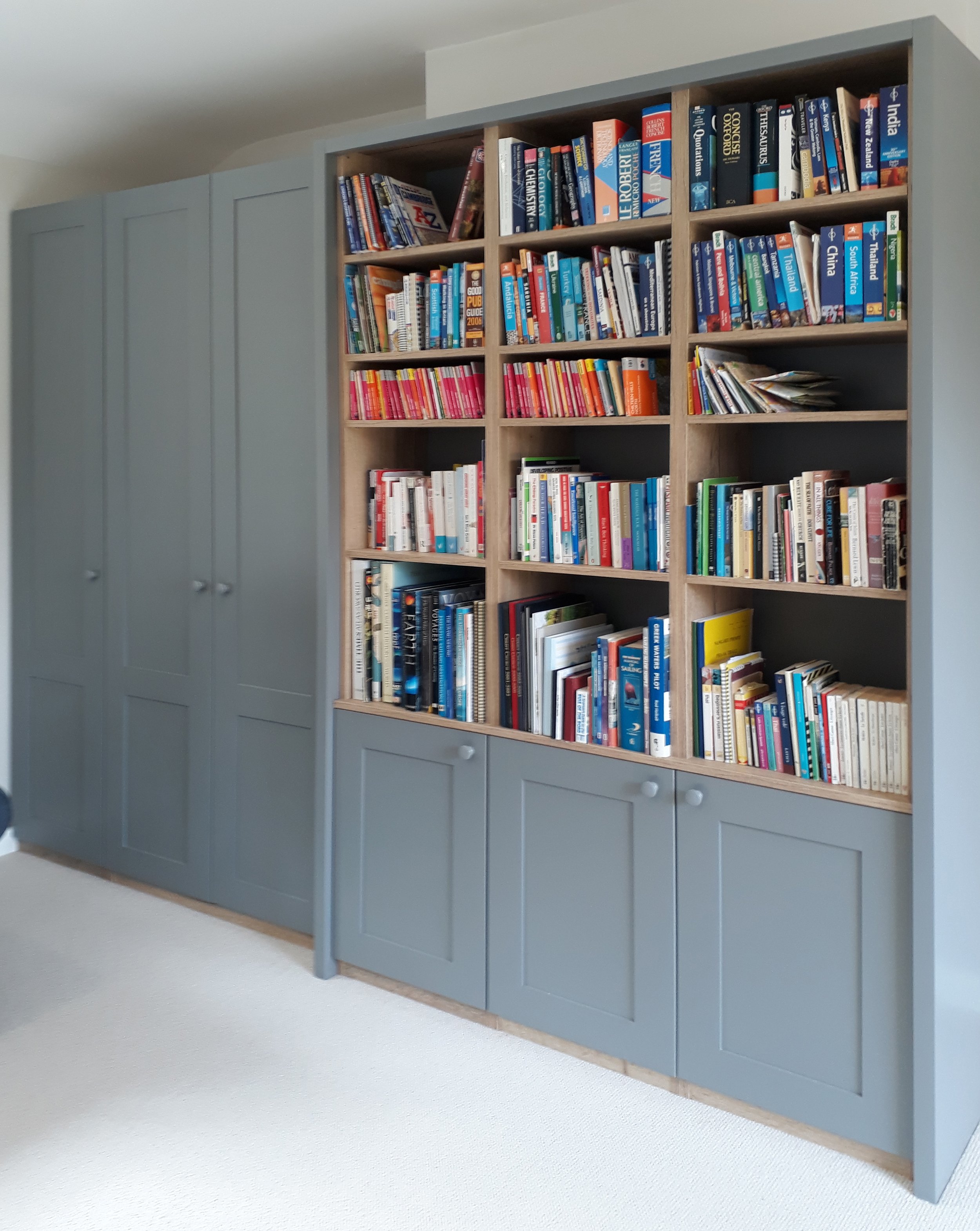 Blue bookshelf with closed cabinet doors at the bottom, filled with books, situated next to grey wall cabinets in a room with beige carpet.