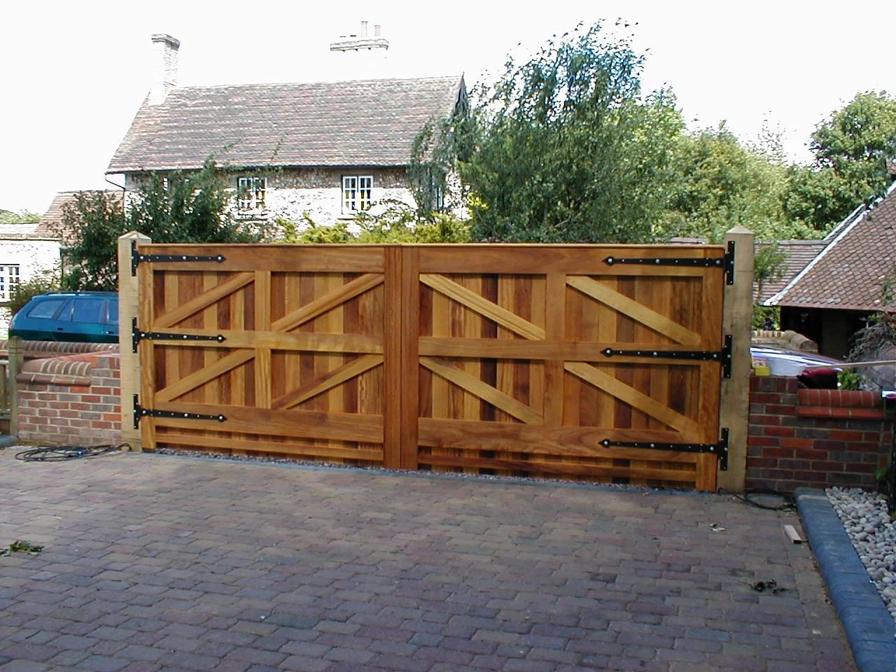 Wooden double gate with black metal hinges in front of a brick driveway, with houses and trees in the background.