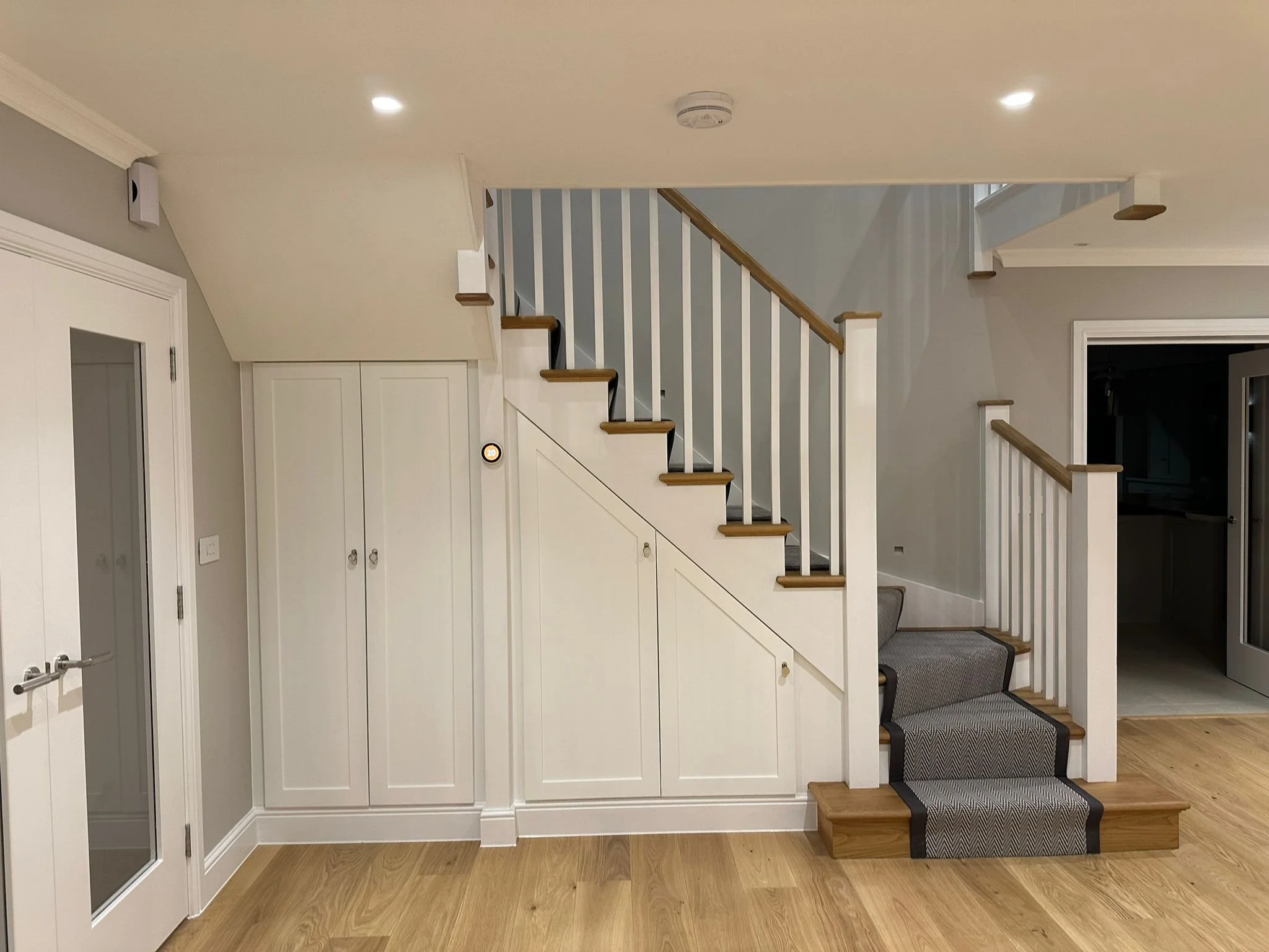 Interior view of a staircase with wooden steps, carpeted with gray, leading upstairs in a house. The wall and railings are painted white, with wooden handrails. There is a closet with double doors under the stairs, and a wooden floor. A door on the l