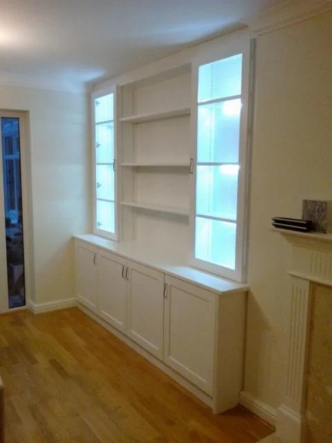 Empty built-in white cabinet with open shelving and closed storage beneath, located next to a fireplace with a decorative mantel and two windows in a room with wooden flooring.