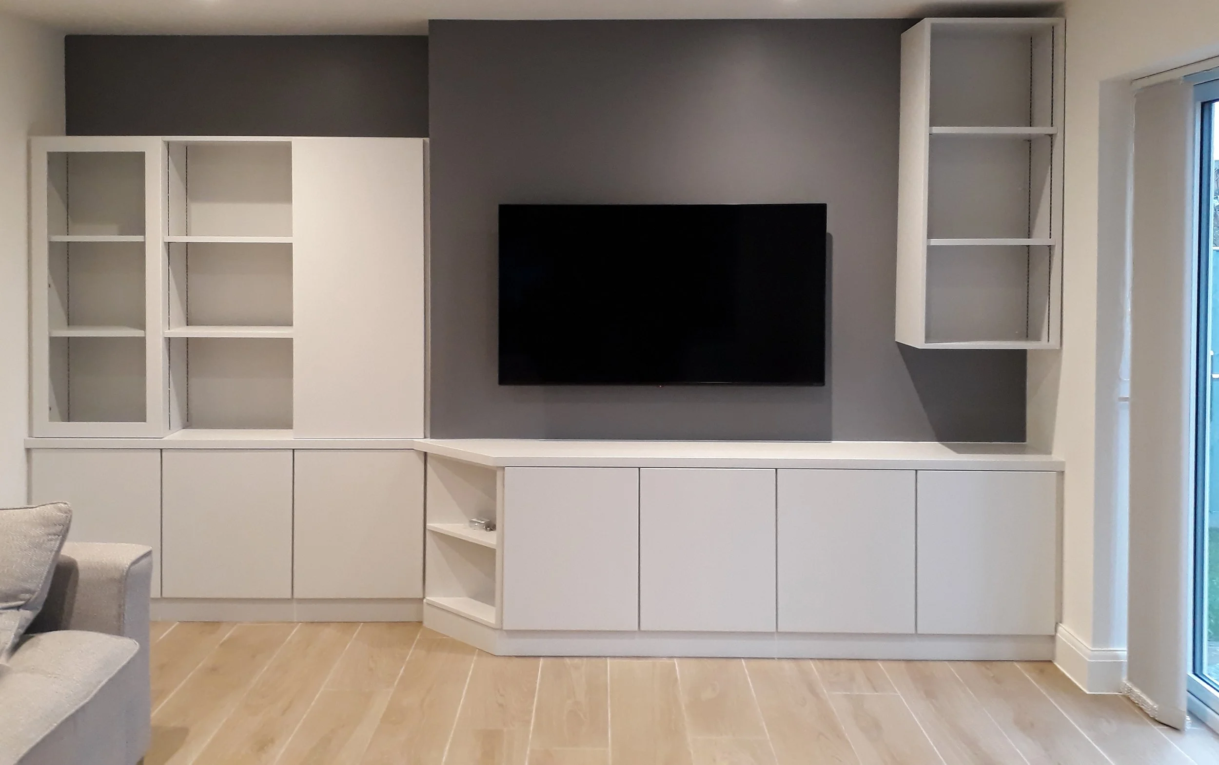 Modern living room TV unit with white cabinetry against a gray wall, featuring open shelves and closed cabinets, a mounted flat-screen TV, and light-colored wood flooring.