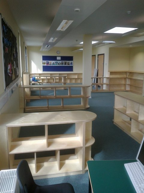 Empty classroom or library with wooden bookshelves, a bulletin board, and chairs. Natural light coming through windows and ceiling lights.
