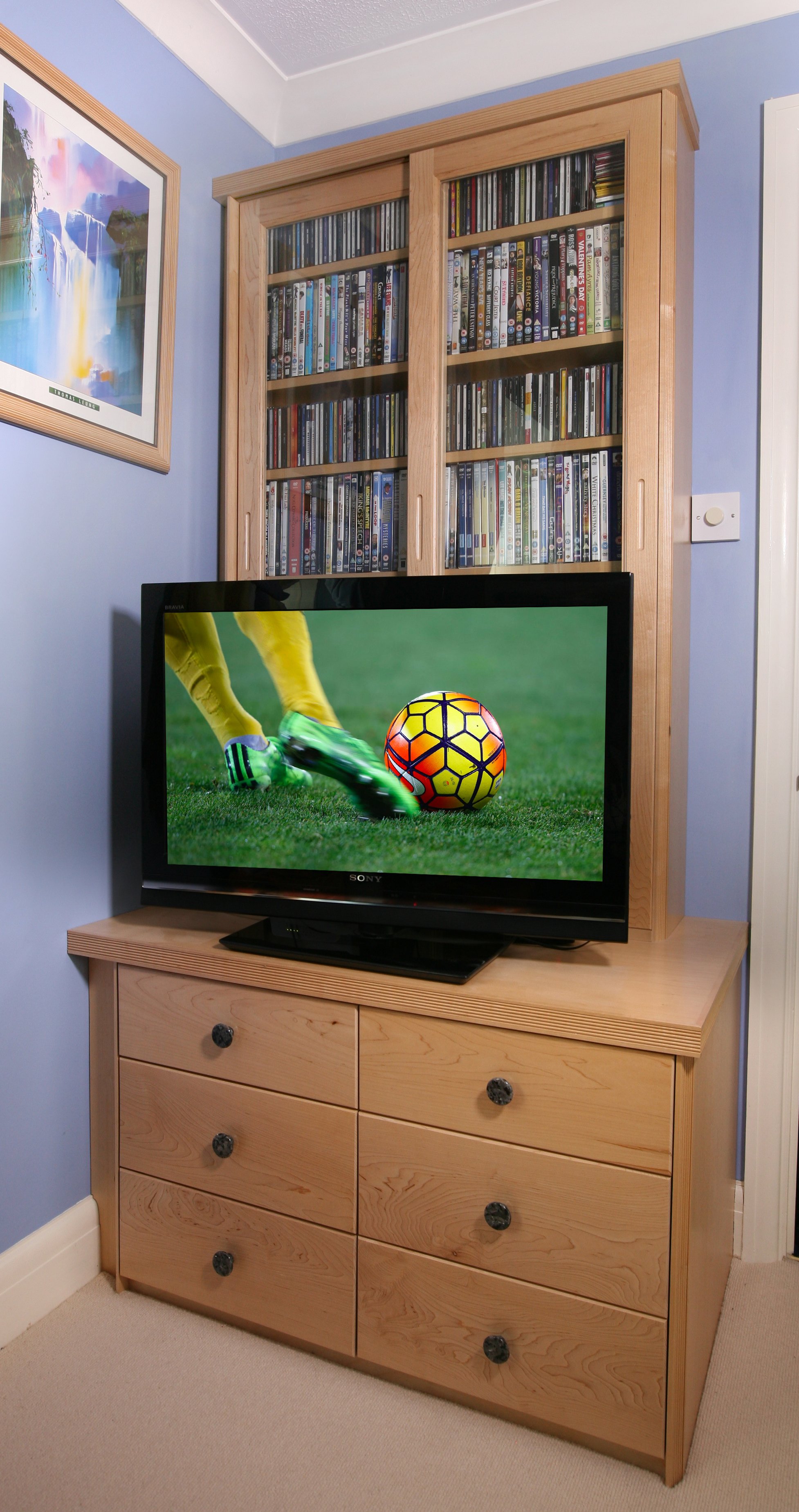 A wooden dresser with six drawers and black knobs, a flat-screen TV on top showing a soccer game, a wooden cabinet filled with DVDs or CDs behind it, a blue wall with a framed painting of a waterfall, and a beige carpeted floor.