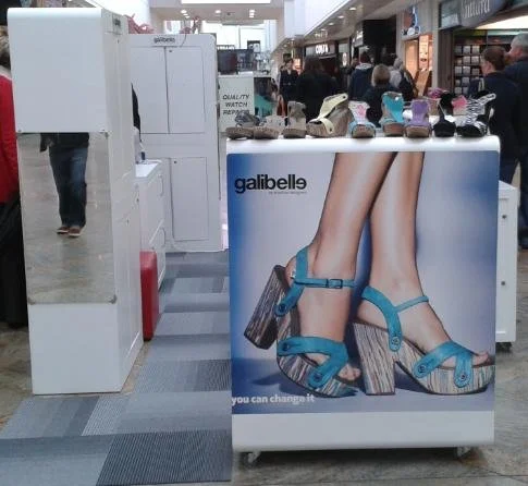 Shoe kiosk and promotional display at a mall, featuring a large image of blue high-heeled sandals for sale.
