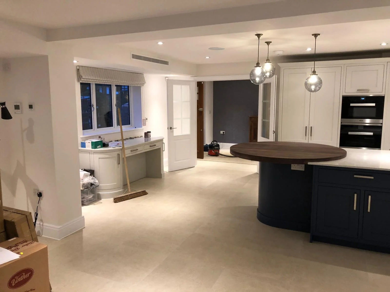 Empty kitchen with white cabinets, gray wall, and three hanging glass pendant lights above a circular dark wood island, with a window, door, and visible appliances.