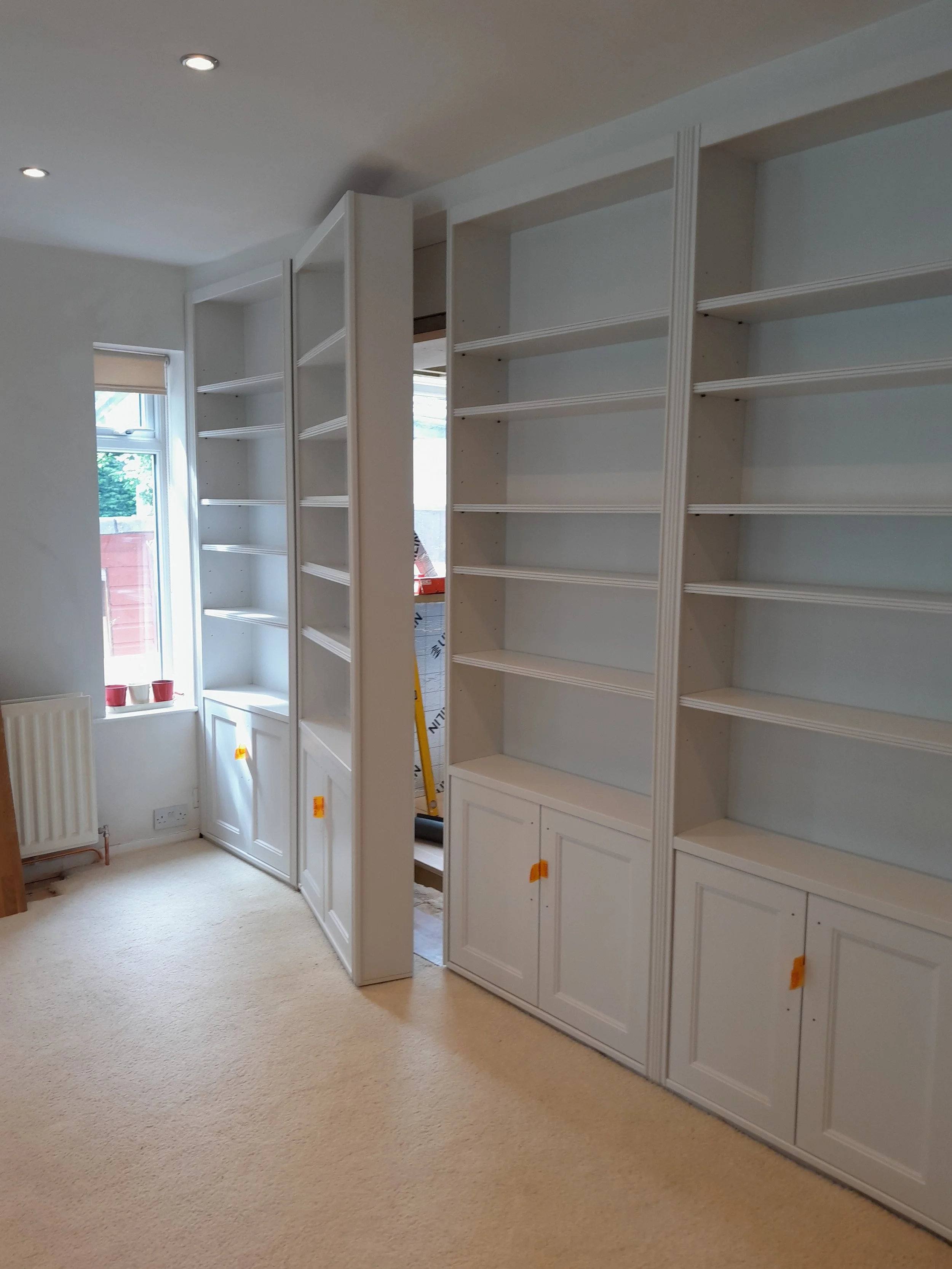 White built-in shelves and cabinets in a room with beige carpet, window with a blinds, and a radiator.