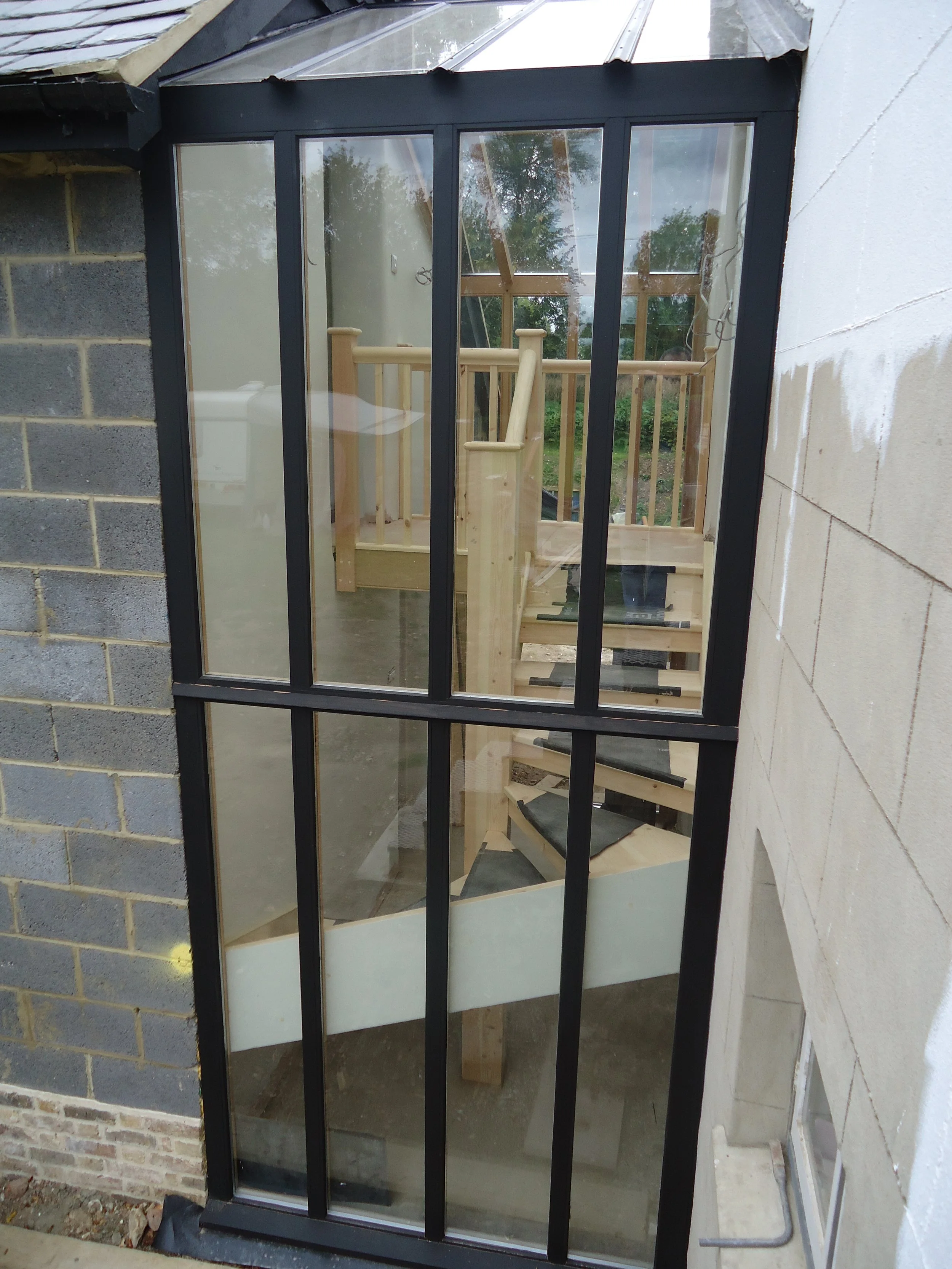 View through a large window showing a wooden staircase and railing inside a building under construction; the exterior includes brick and concrete wall.