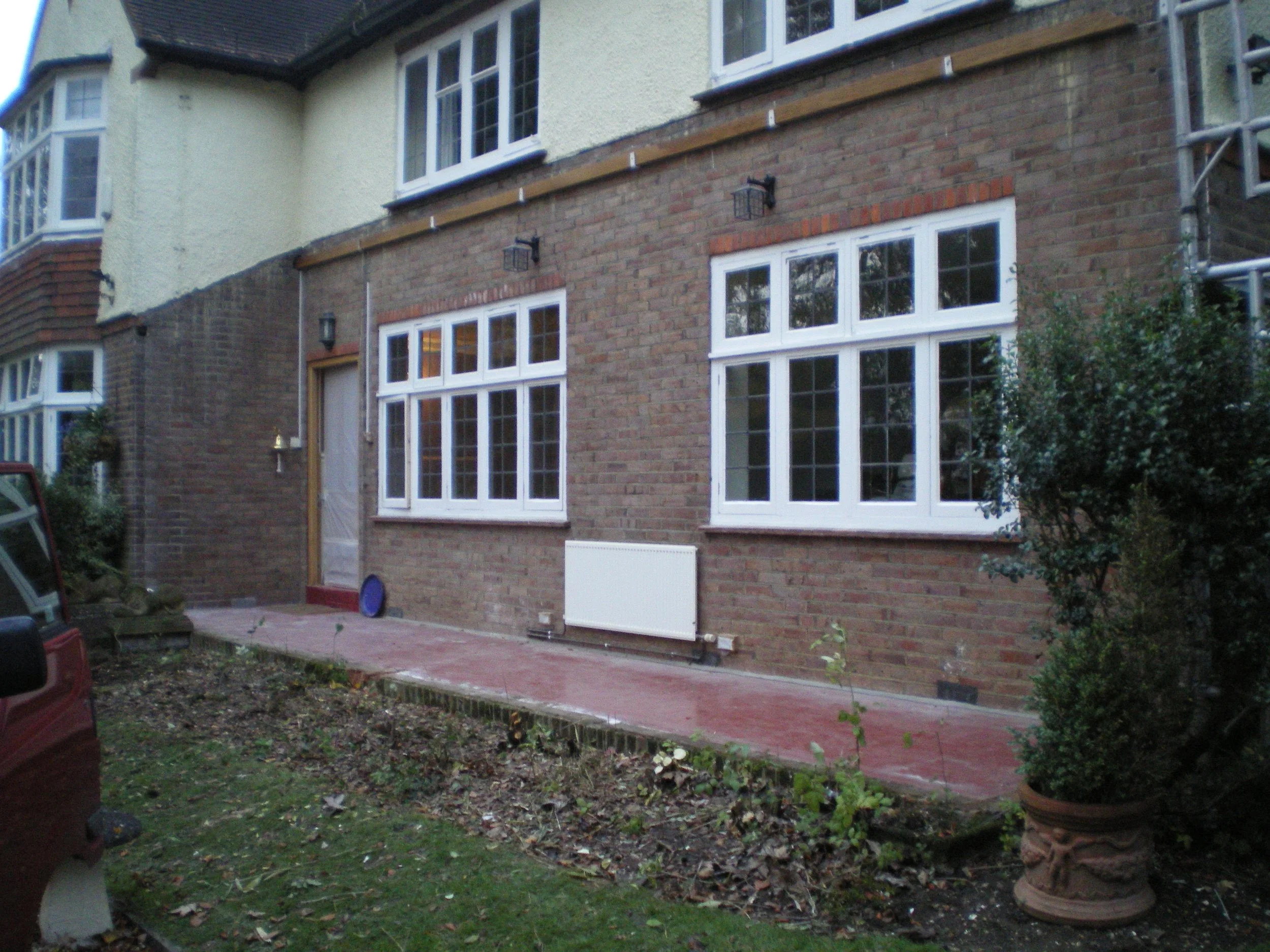Backyard patio area with brick and beige exterior wall, white-framed windows, a door, a radiator, and potted plants, with some gardening tools and a parked vehicle visible.