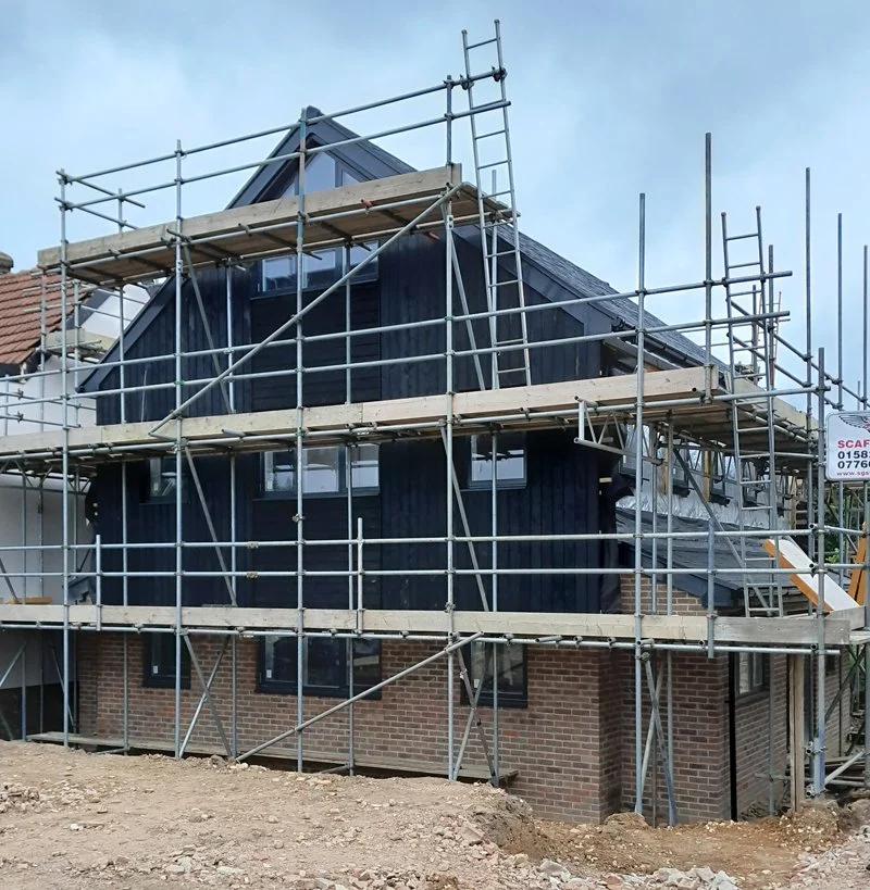 A house under construction with scaffolding around it, featuring a black exterior and a steep roof.