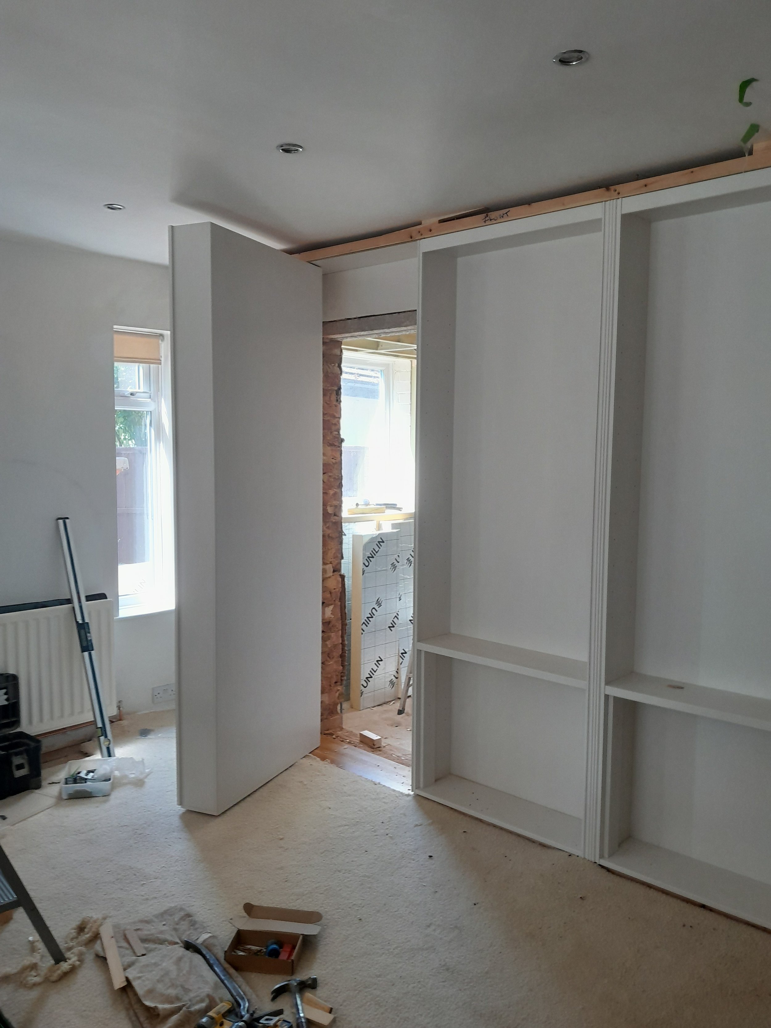 Living room under renovation with construction tools on beige carpet, white built-in shelves, partially built doorway, and unfinished ceiling.