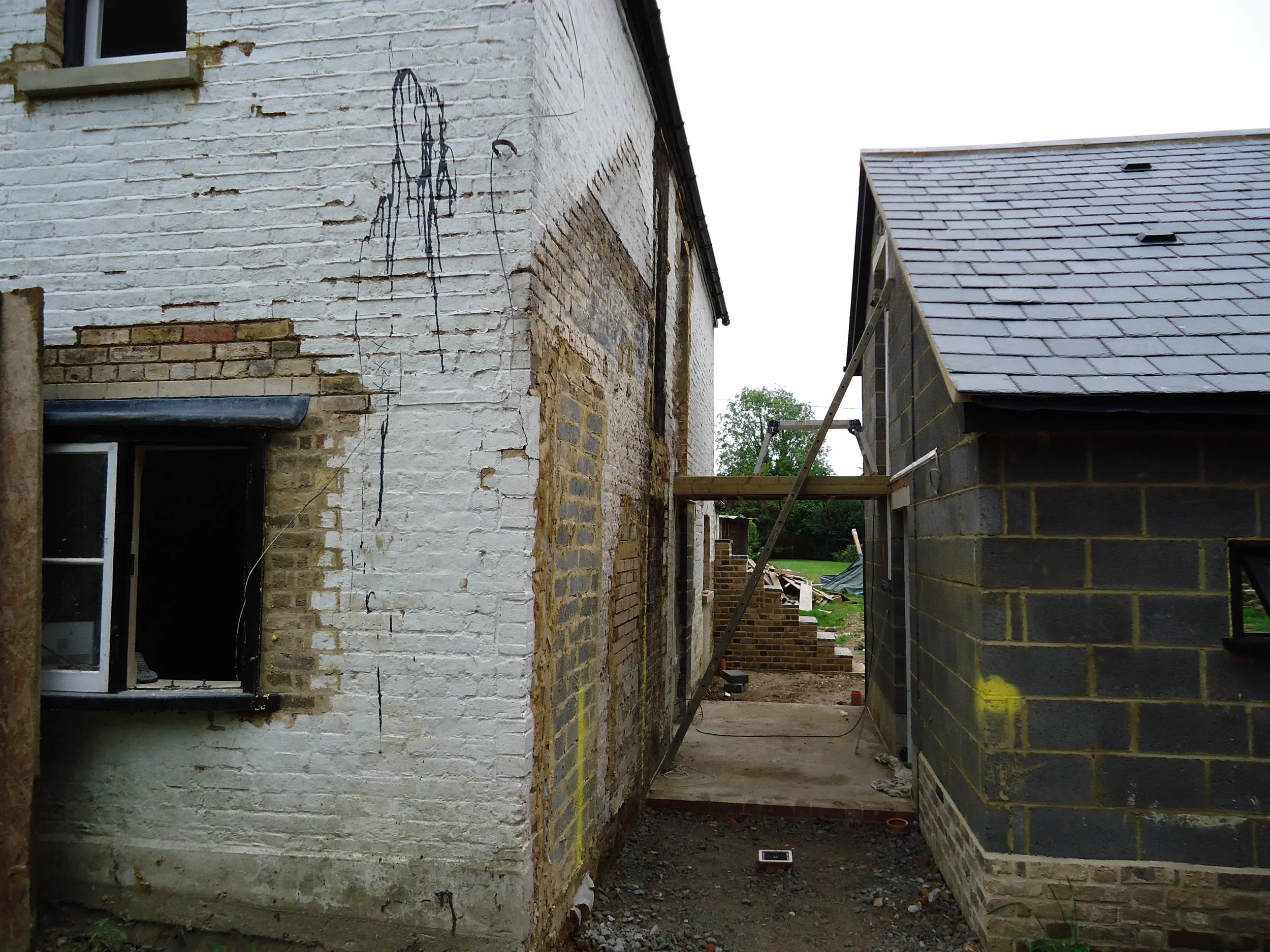 A construction site with two buildings, one painted white with exposed brick and a window, and a smaller gray brick building with a sloped roof, linked by a wooden plank walkway under construction.