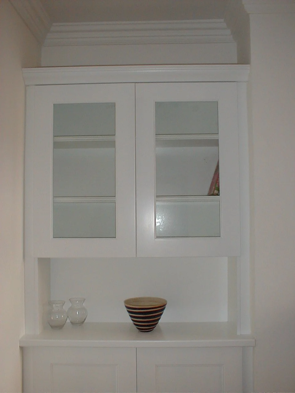 White display cabinet with glass-paneled doors and decorative molding, with vases and a striped bowl on the lower shelf.
