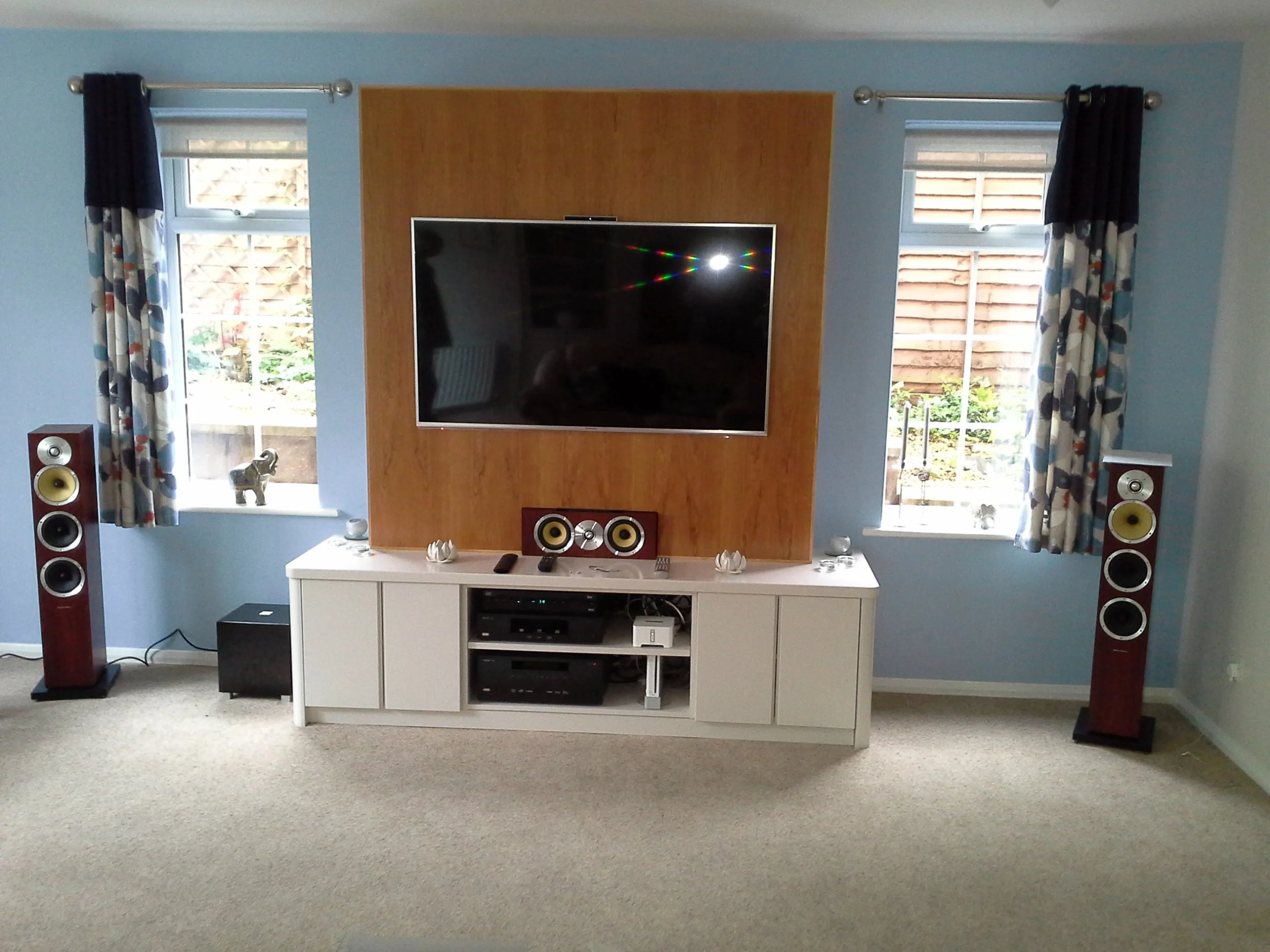 Living room entertainment setup with two windows, a large flat-screen TV on a wooden panel, white TV stand with speakers and electronic devices, and two tall floor-standing speakers on either side, with decorative curtains and a carpeted floor.