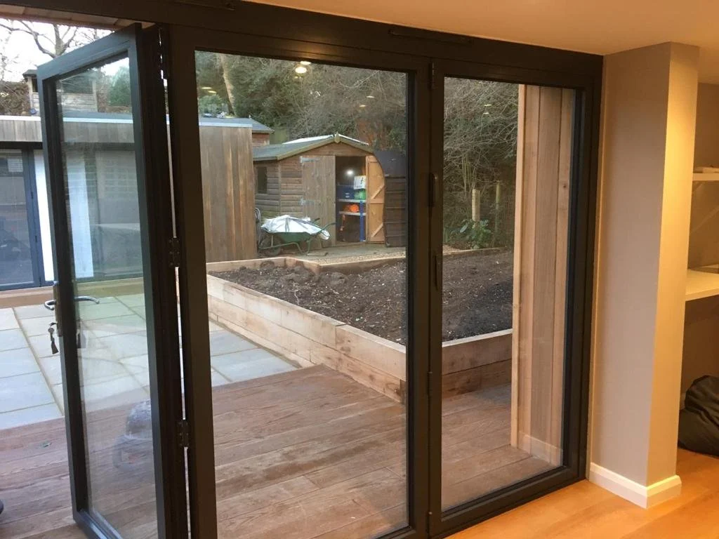 View of a garden area through a large sliding glass door, with a wooden deck in the foreground, a garden shed, and some gardening tools outside.