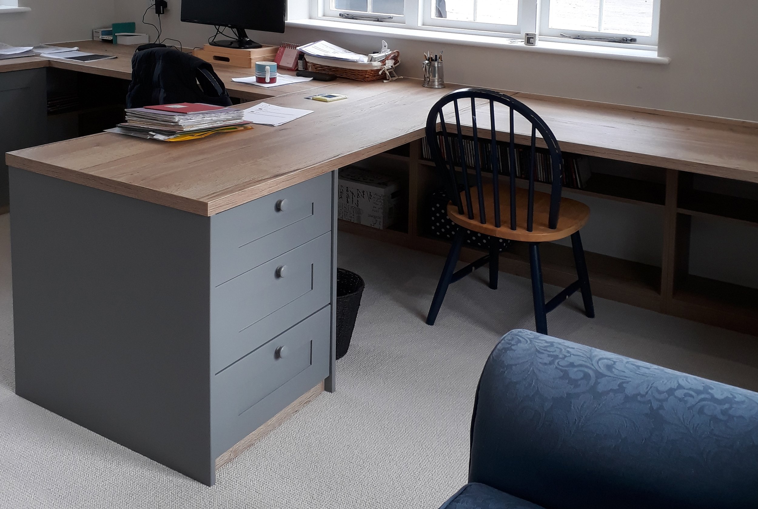 Office workspace with a large wooden desk, blue filing cabinet with four drawers, black wooden chair with polka-dotted cushion, shelves with books, papers, and office supplies, and a window with natural light.