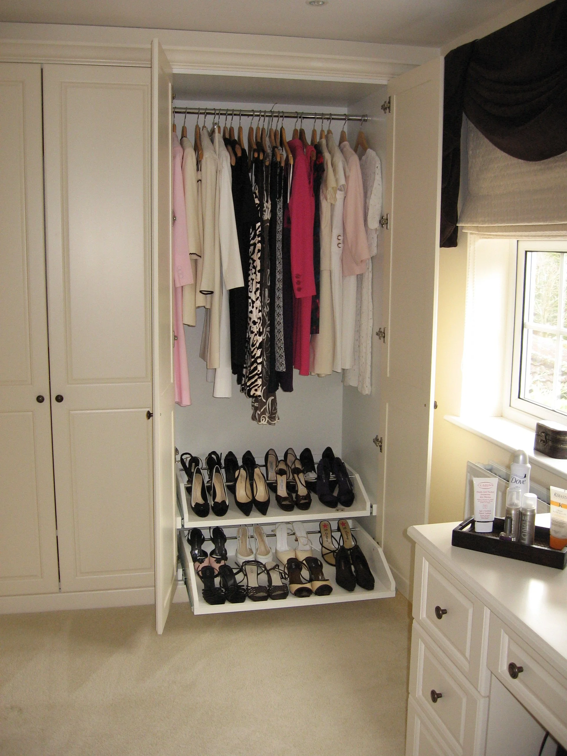 Open closet with hanging clothes and a shoe rack containing high heels, sandals, and flats, next to a window with a black-and-white curtain and a dresser with toiletries.
