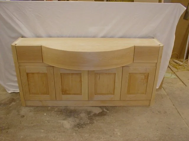 Unfinished wooden cabinet with paneled doors and a curved top section in a workshop.