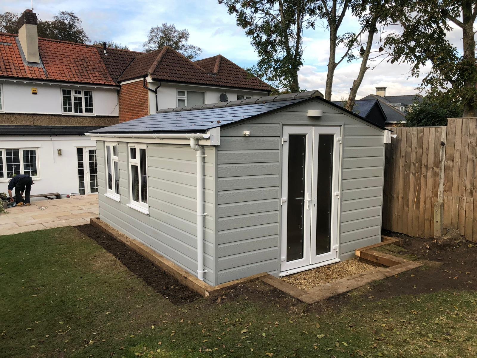 A small gray garden shed with a double glass door, situated on a plot with some brick and gravel edging, with houses and trees in the background.