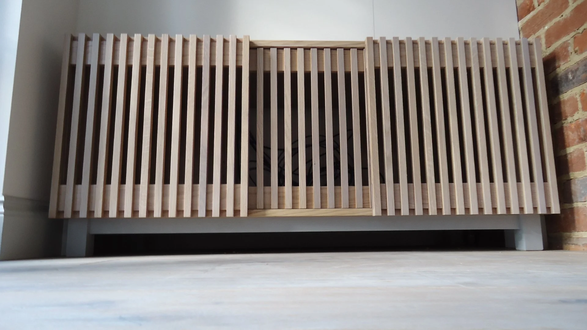 A wooden slatted furniture piece, possibly a bench or a storage unit, with a rectangular shape and vertical slats, placed against a brick wall and a white wall, with a light-colored floor beneath.