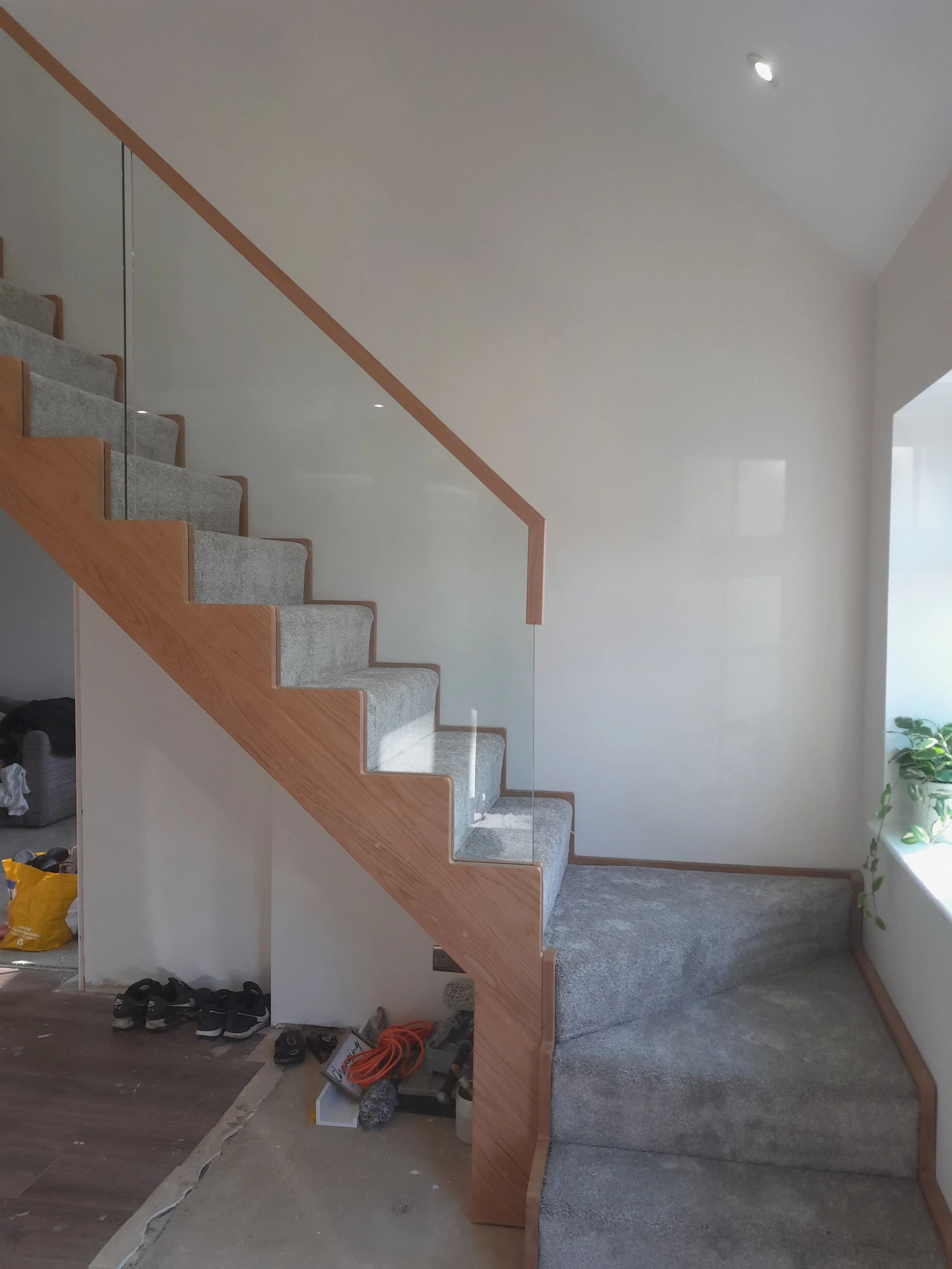 Interior view of a modern staircase with wooden handrails, gray carpeted steps, and a glass panel balustrade, located next to a window with a potted plant.