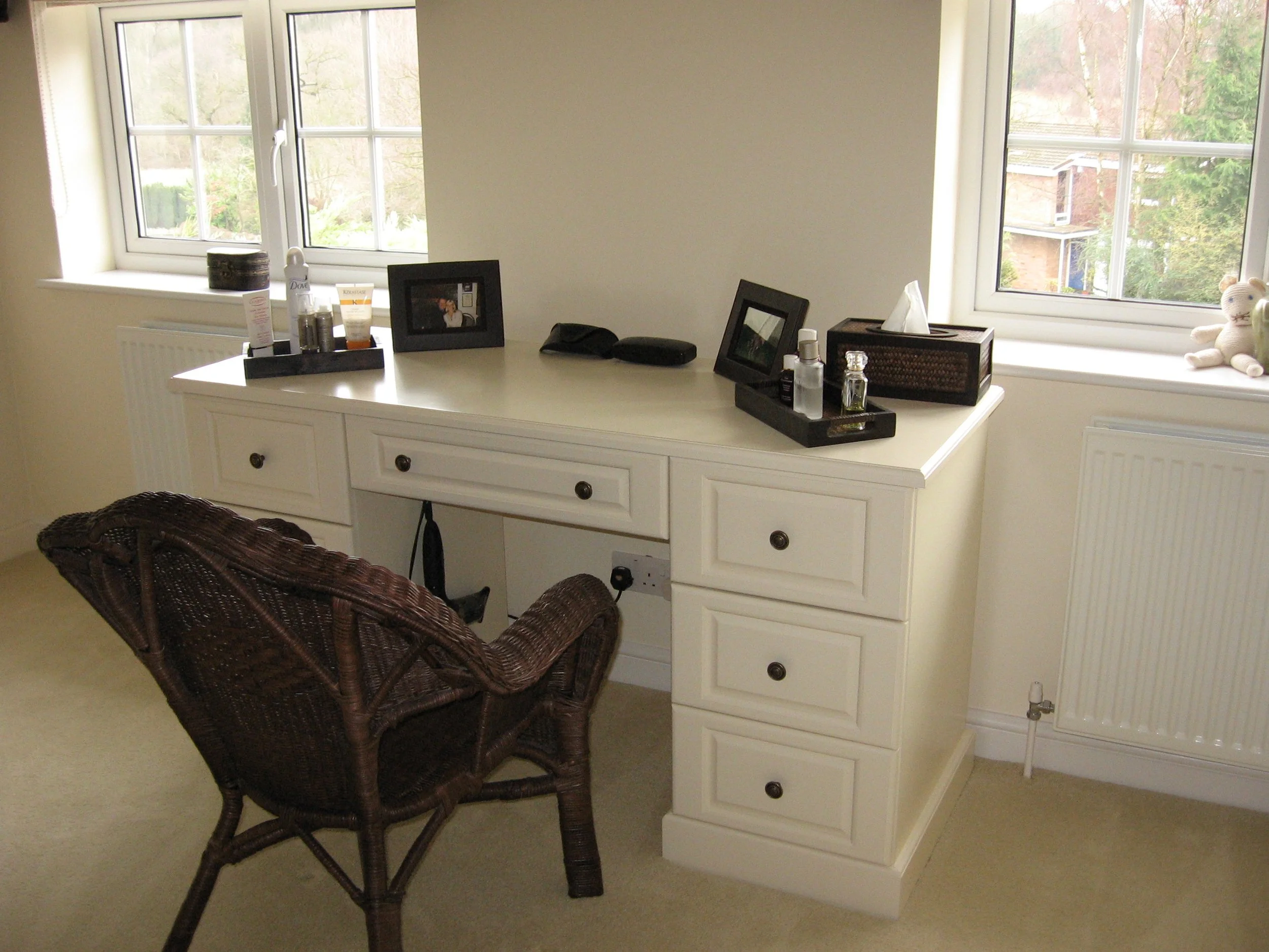 A white desk with eight drawers, placed under two windows, with various personal items and photos on top, and a brown wicker chair in front of it inside a room with beige walls and carpet.