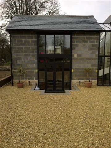 Small brick building with large glass double doors and windows, surrounded by a gravel yard and potted plants.