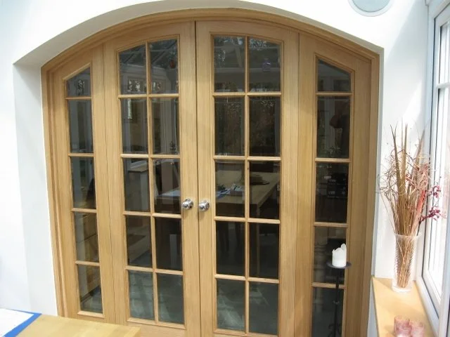 Double wooden framed glass doors with an arched top leading to another room, with a small side table holding a candle and decorative branches next to a window.