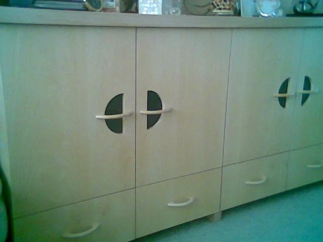 A beige wooden sideboard with cutout handles and two doors in the center, set against a light-colored wall with various decorative items on top.