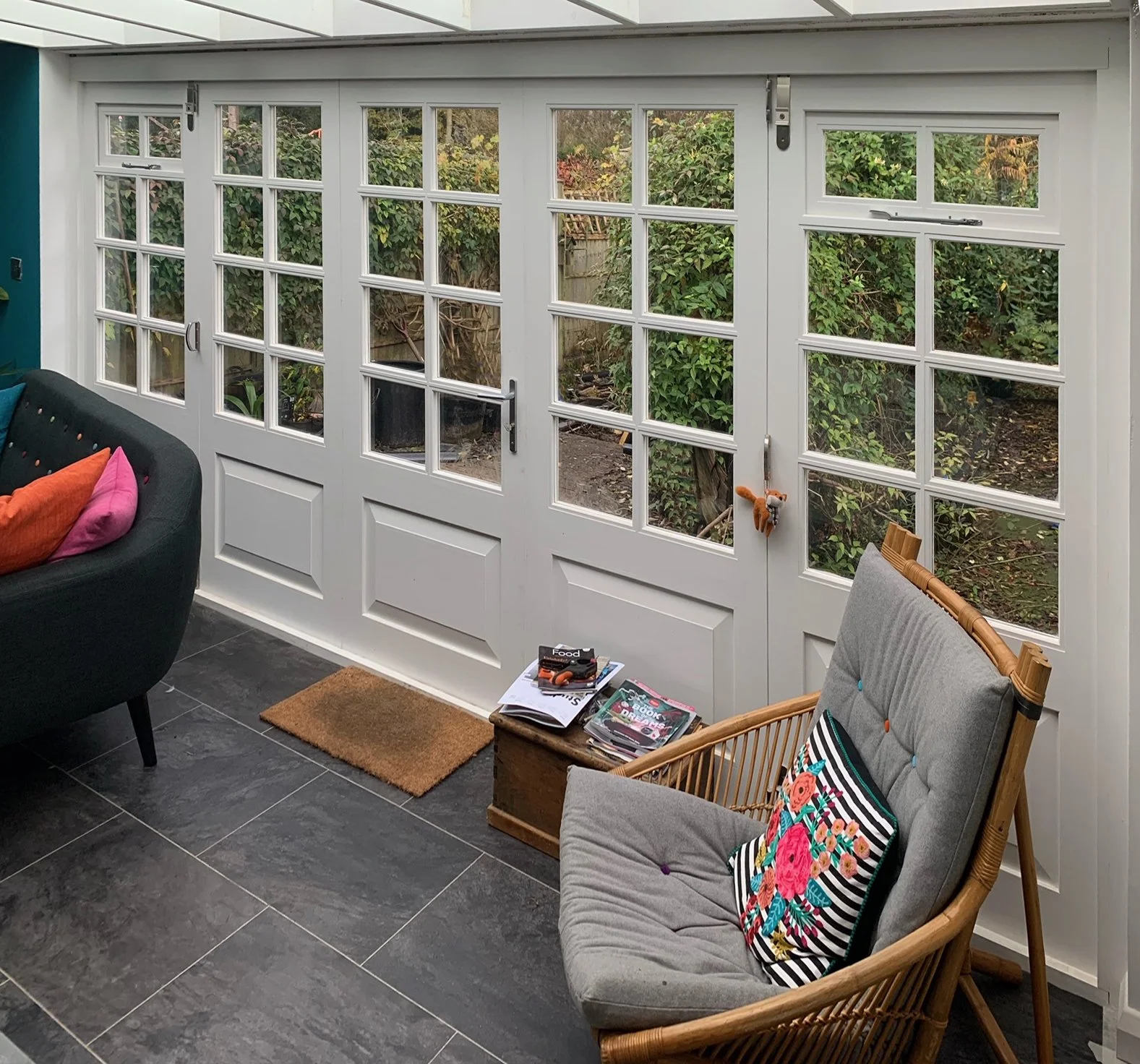 Indoor sitting area with large white-paneled glass doors looking to a garden, a gray cushioned lounge chair with a colorful floral pillow, a small corner table with stacked magazines, a black sofa with multi-colored pillows, and a doormat on grey til