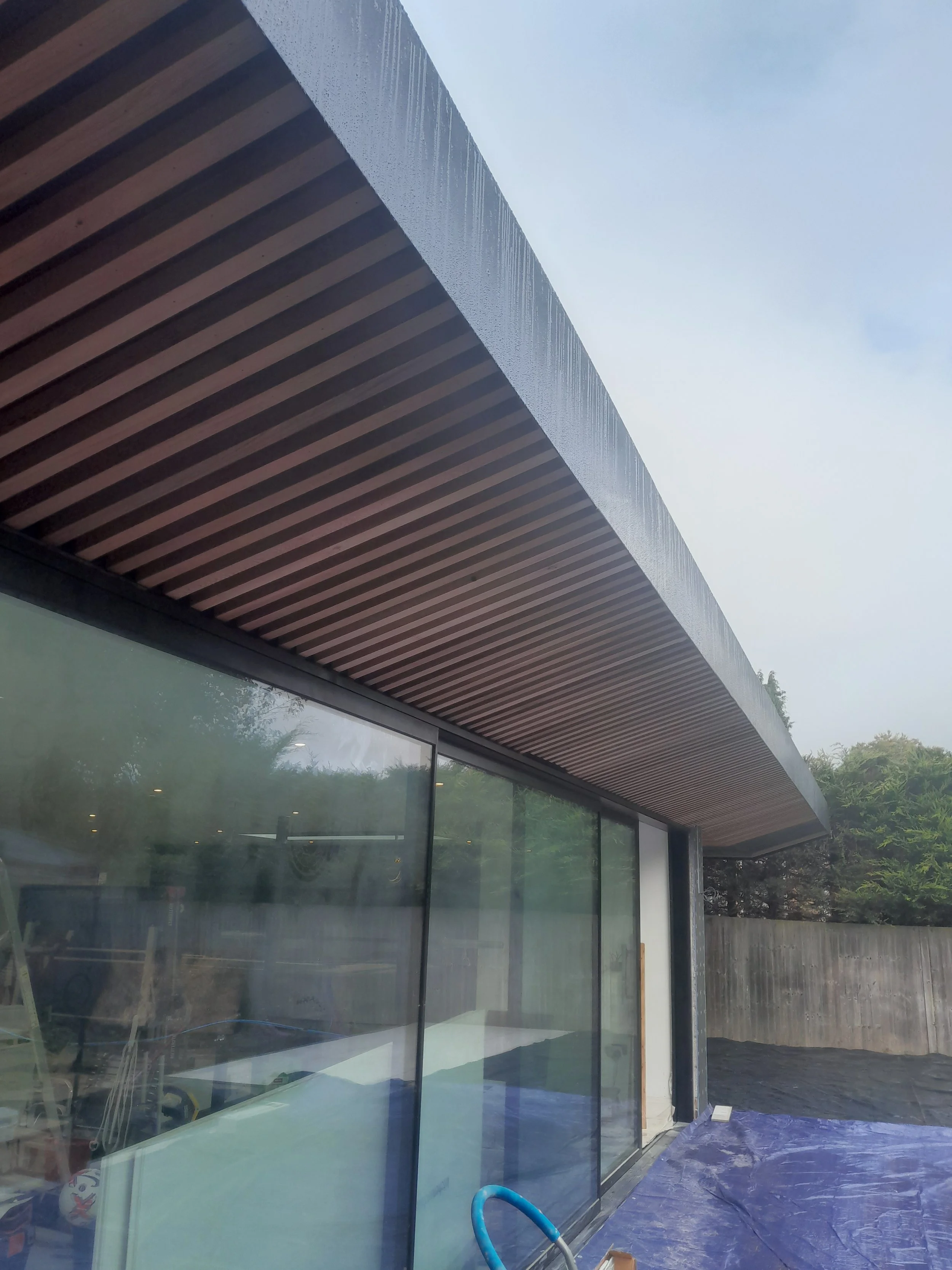 Modern house with large glass windows, wooden overhang, and a patio covered with blue tarp, surrounded by trees and a wooden fence.