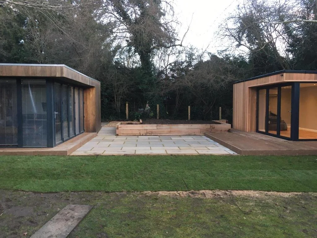A modern backyard with two wooden houses featuring large glass sliding doors, a paved patio area, a grassy lawn, and a wooded background.