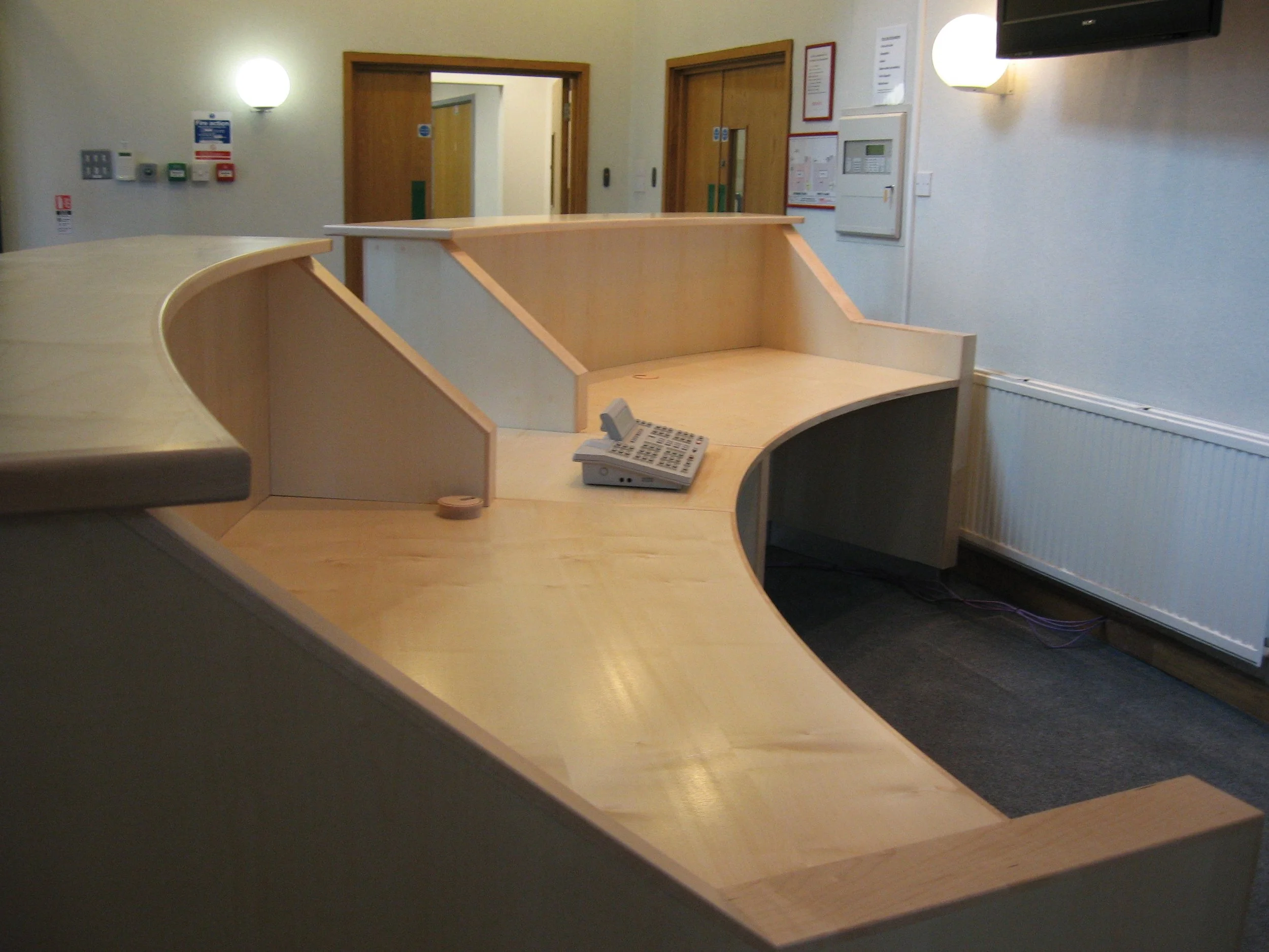 Empty reception desk with a landline telephone, in a room with wooden doors and various control panels on the wall.