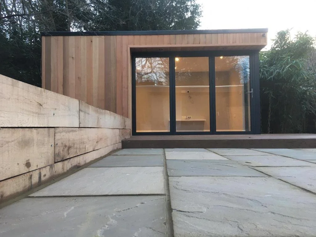 View of a modern small wooden building with large glass doors, situated outdoors on a stone patio, with natural greenery in the background.