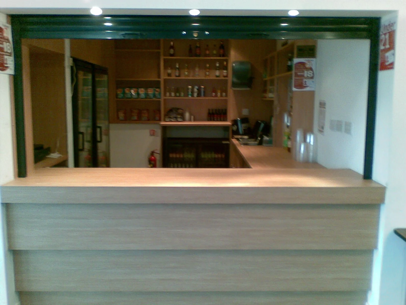 View of a small snack or convenience store counter with a wooden front, behind which are shelves stocked with canned and bottled goods, a refrigerator with glass doors, and a cash register area with two computers.