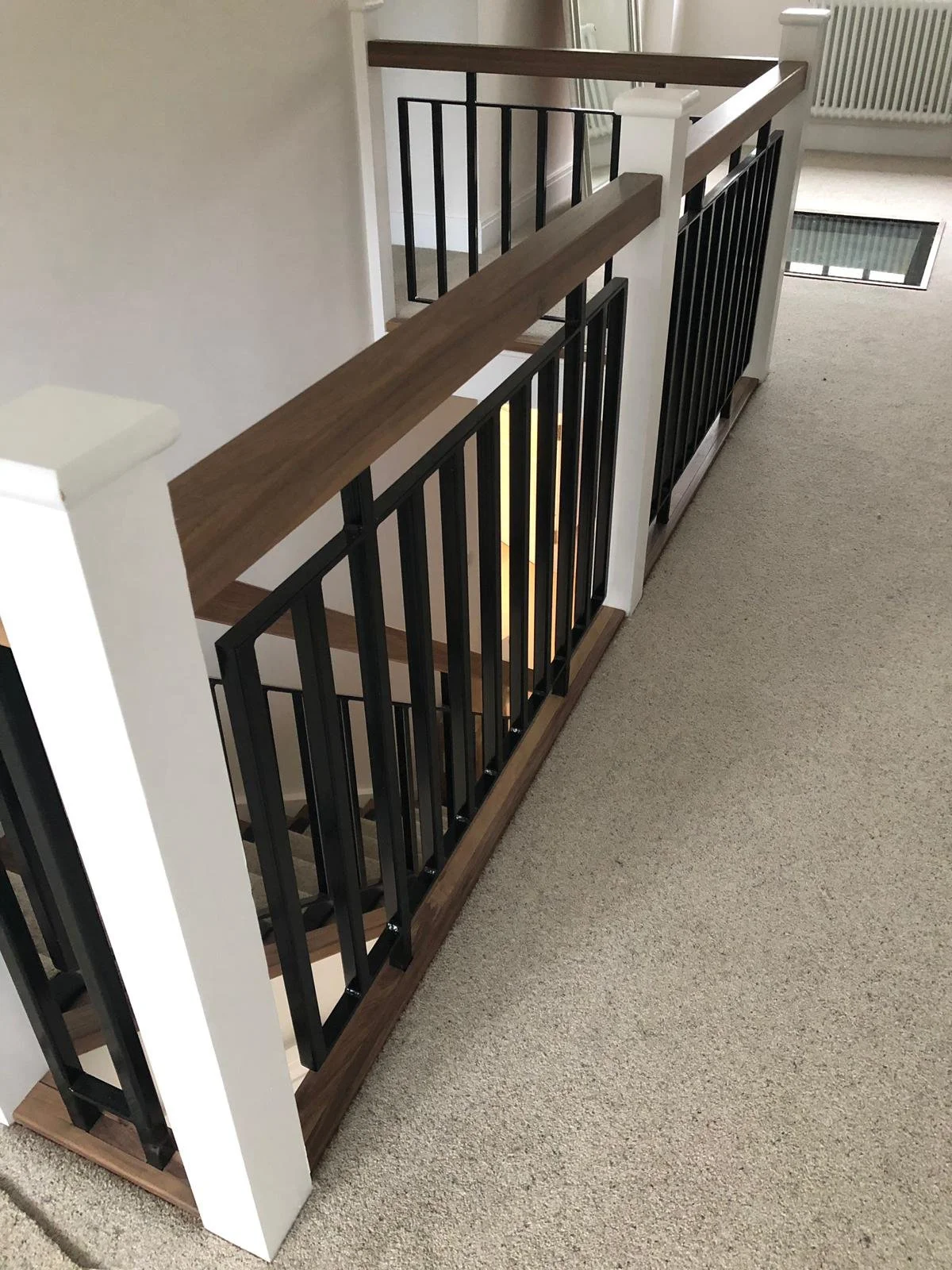Top view of a staircase landing with a black metal railing, wooden handrail, and beige carpeted floor.