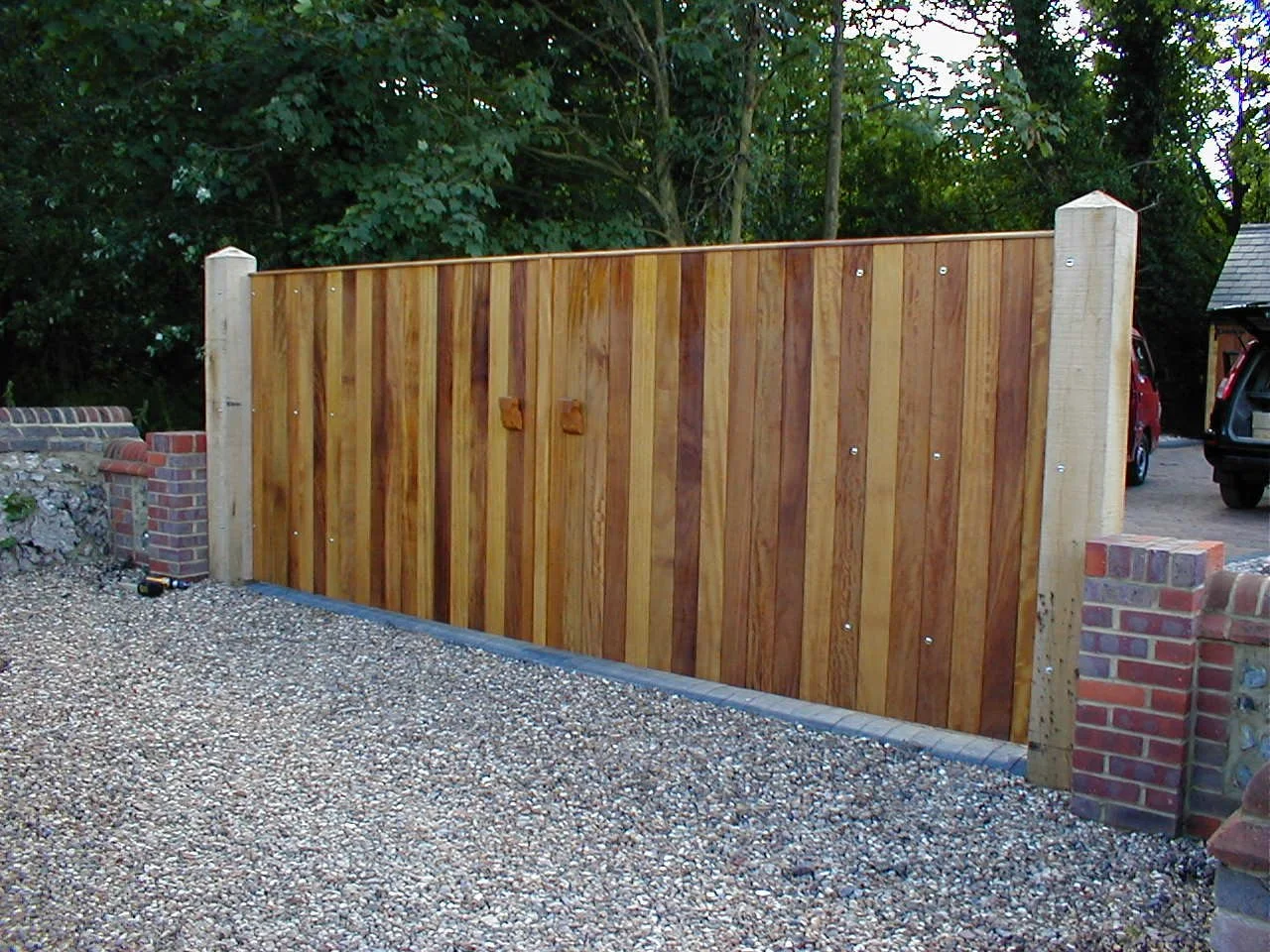 Wooden privacy fence with brick pillars on either side, installed on a gravel surface, with trees and cars visible in the background.