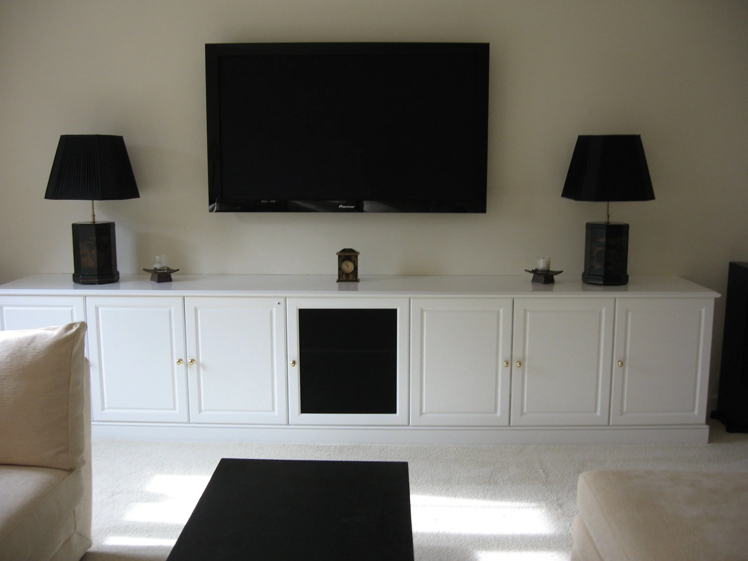 Living room entertainment center with a mounted flat-screen TV, two black table lamps, a small clock, and two small decorative trays on a white cabinet with gold knobs. Beige sofas are partially visible.