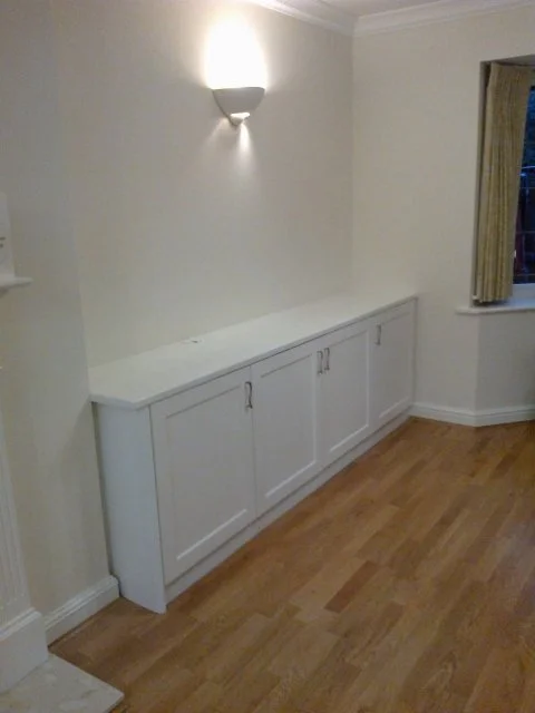 Empty white cabinet with doors under a wall-mounted light fixture in a room with hardwood floors and a window with curtains.