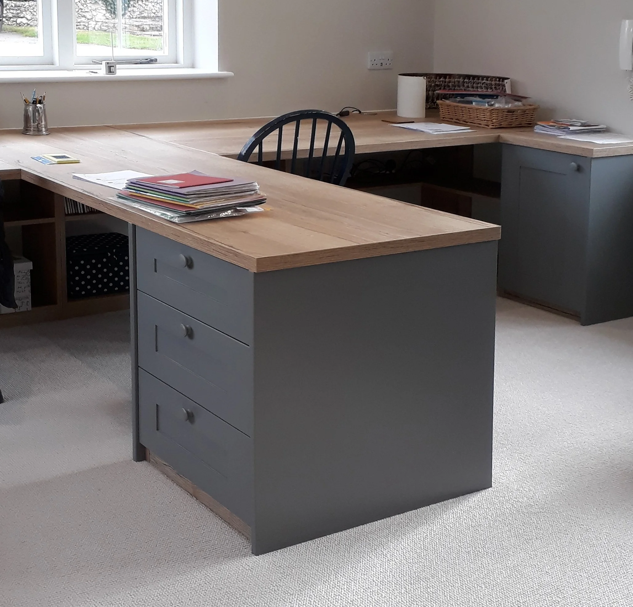 Home office with L-shaped wooden desk, gray lower cabinets, black chair, window, and various office supplies on the desk.