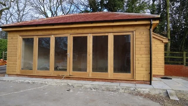 A wooden garden shed with a red roof, large glass doors, and windows, situated on a concrete patio outdoors.