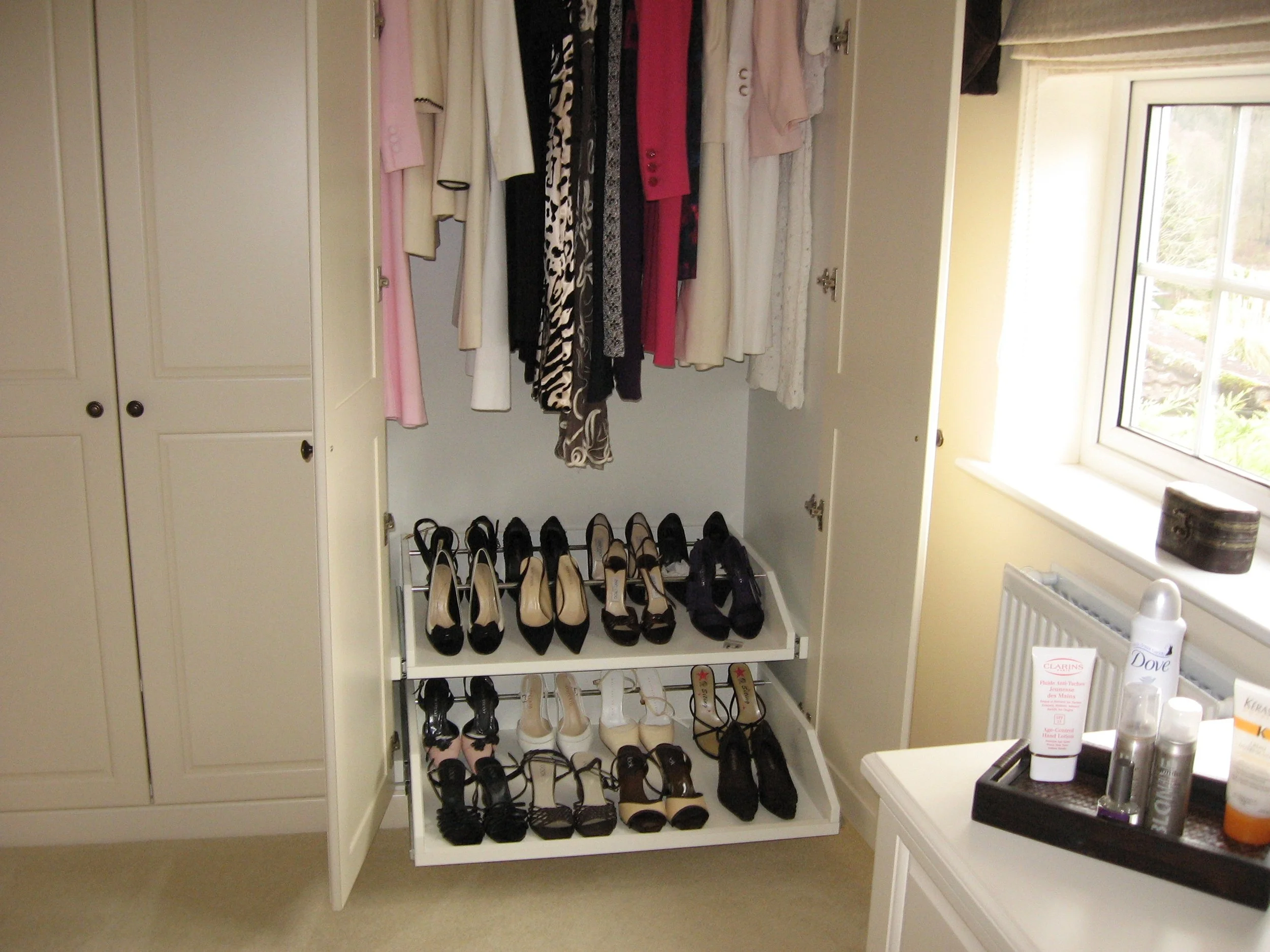 A closet with white shelves and double doors, containing hanging dresses and high-heeled shoes stored on built-in racks, with a window and a small table holding toiletries nearby.