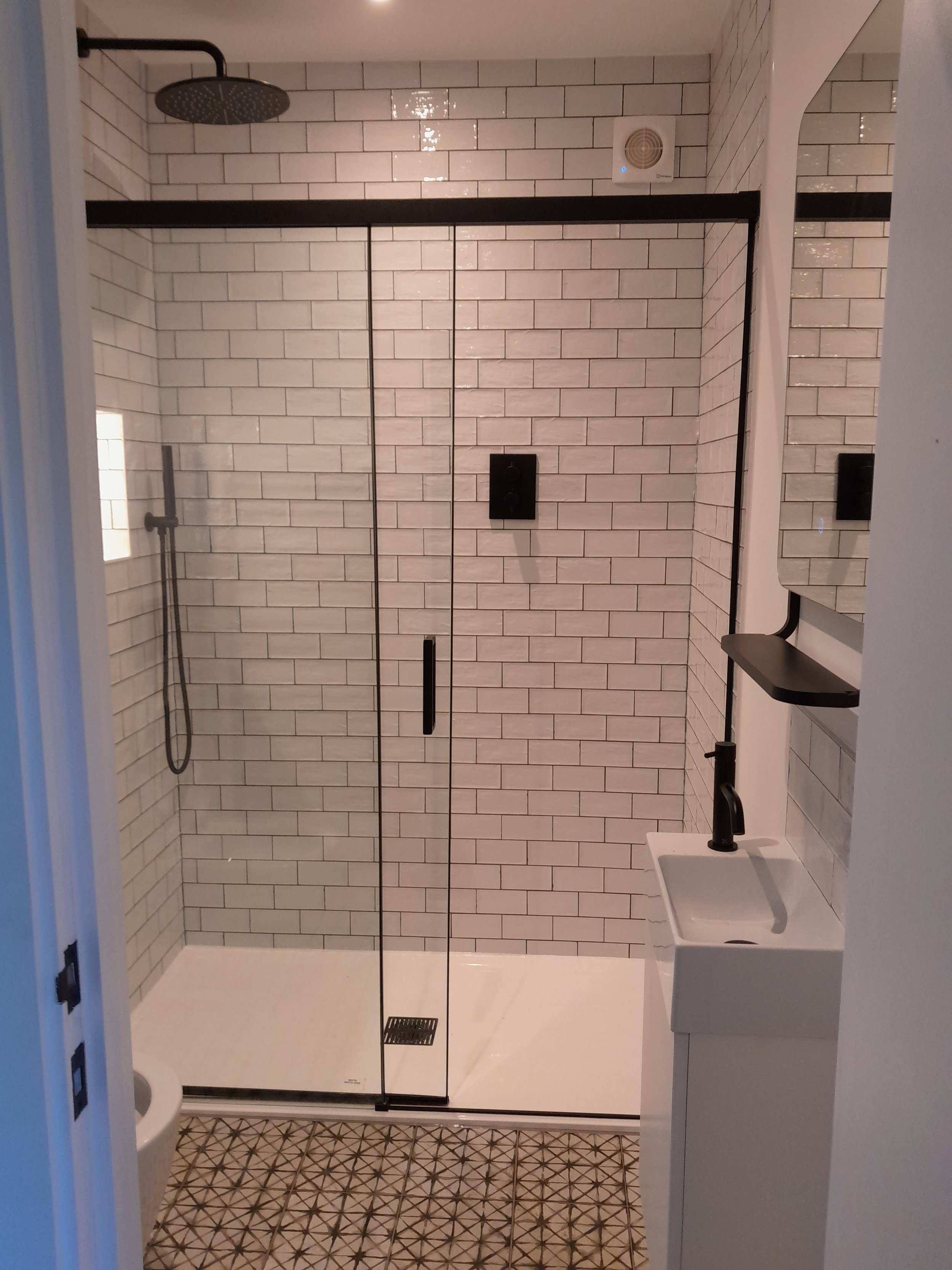 Modern bathroom with glass-enclosed shower, white subway tile walls, a small white sink with a black faucet, a mirror, and patterned floor tiles.