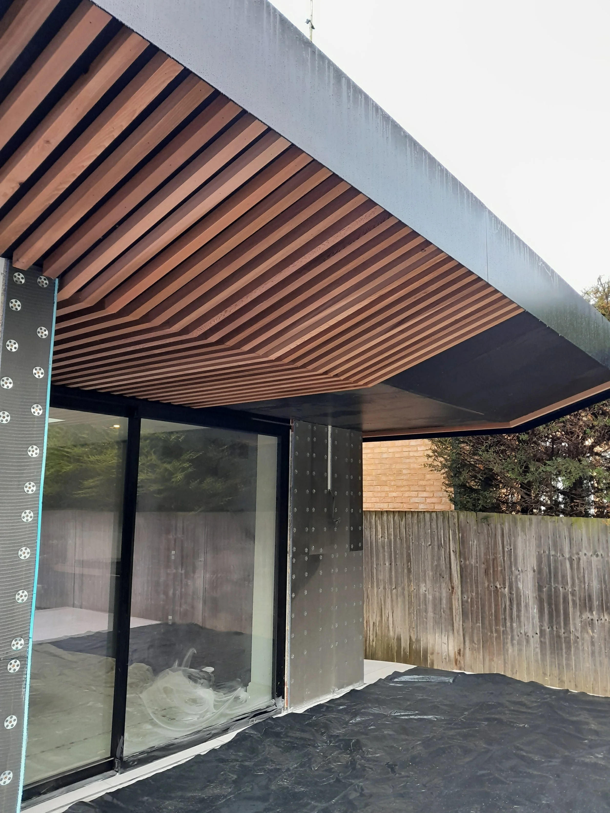 Building under construction with wooden ceiling panels and exterior metal and glass walls, a wooden fence in the background, and construction materials on the ground.