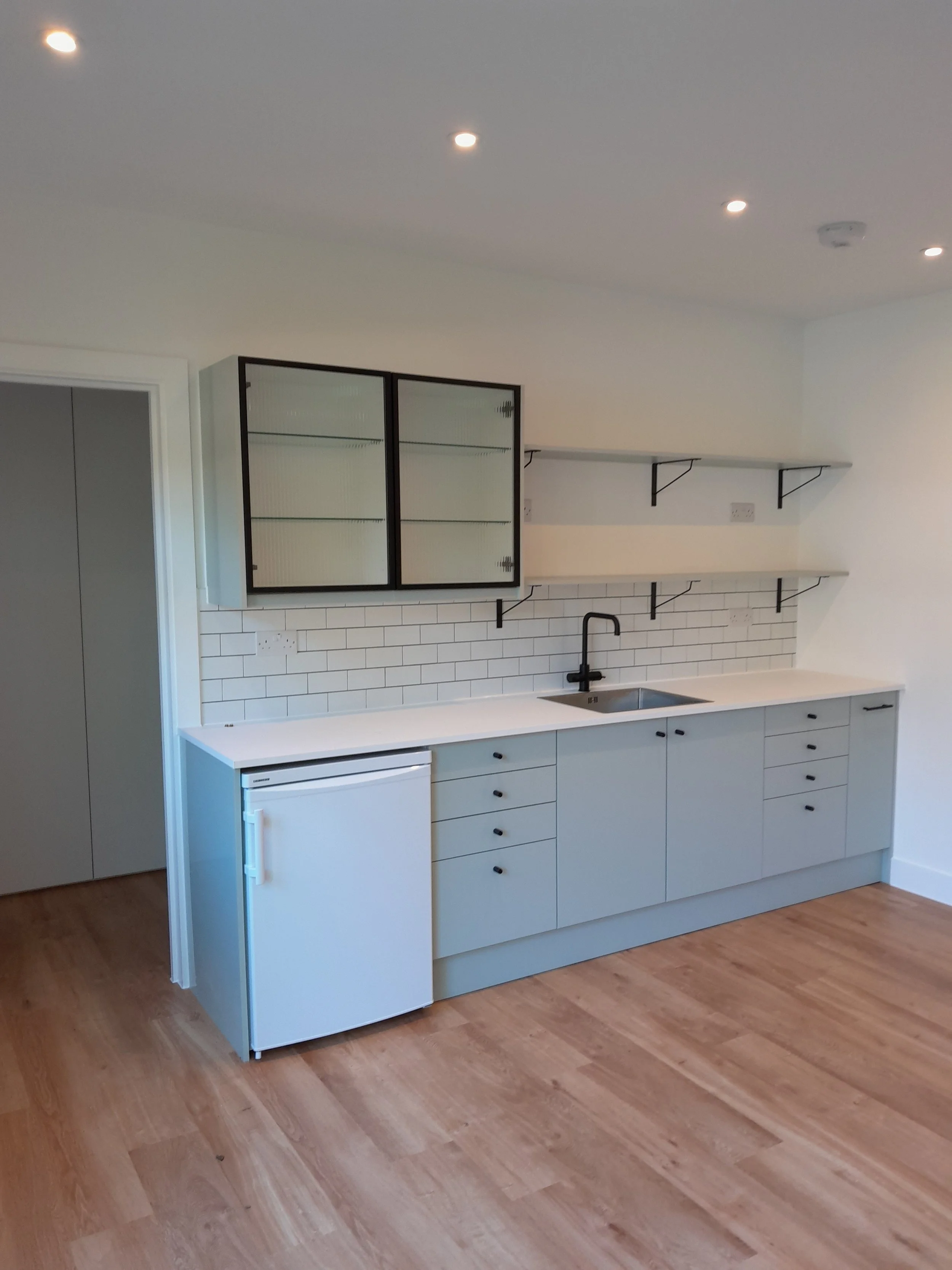 Minimalist kitchen with white cabinets, black hardware, a small white refrigerator, open shelves, a black faucet, and a white tile backsplash.